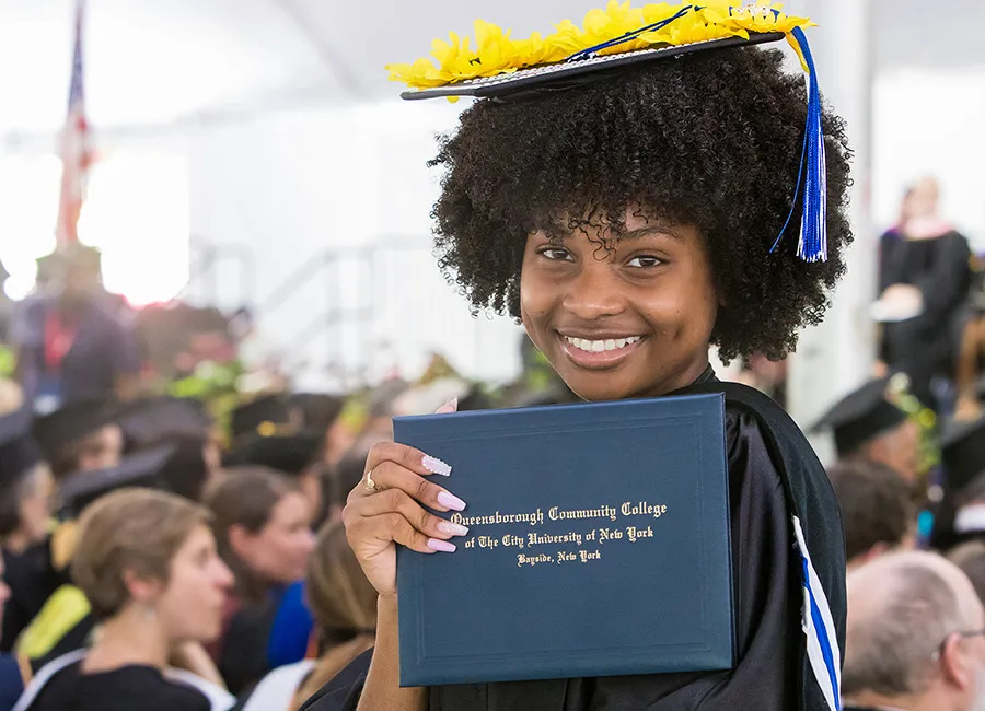 A woman in a graduation cap holds up a diploma from Queensborough Community College