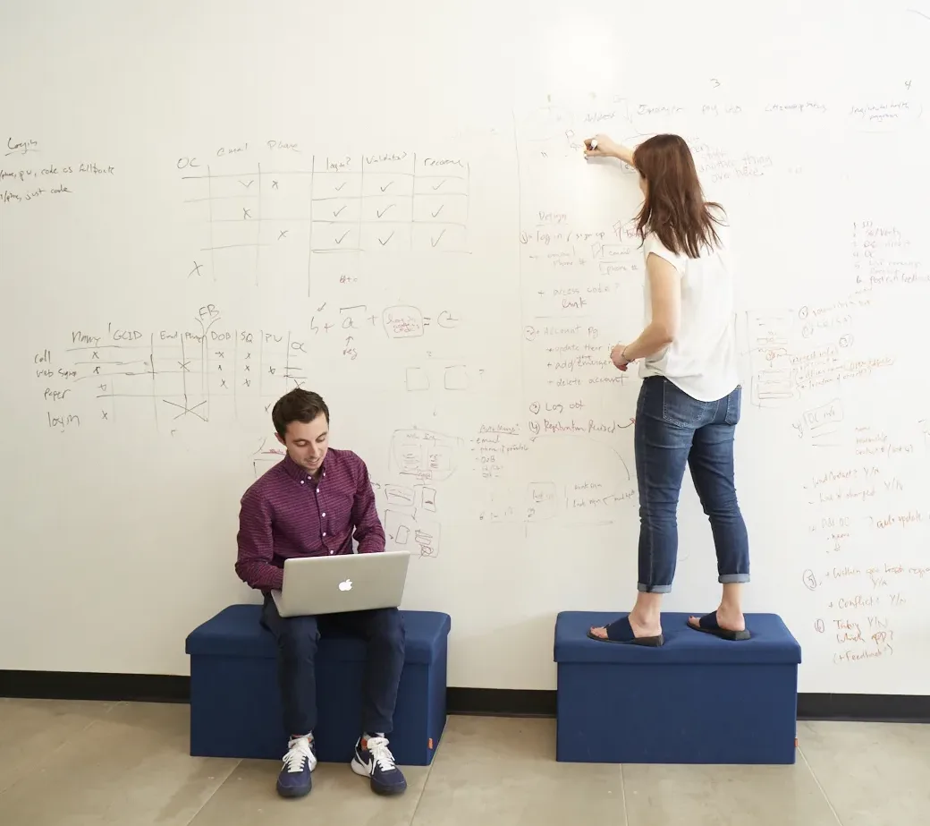 Two people on ottomans, one sitting and typing on a laptop, and the other writing notes and equations on a whiteboard wall.