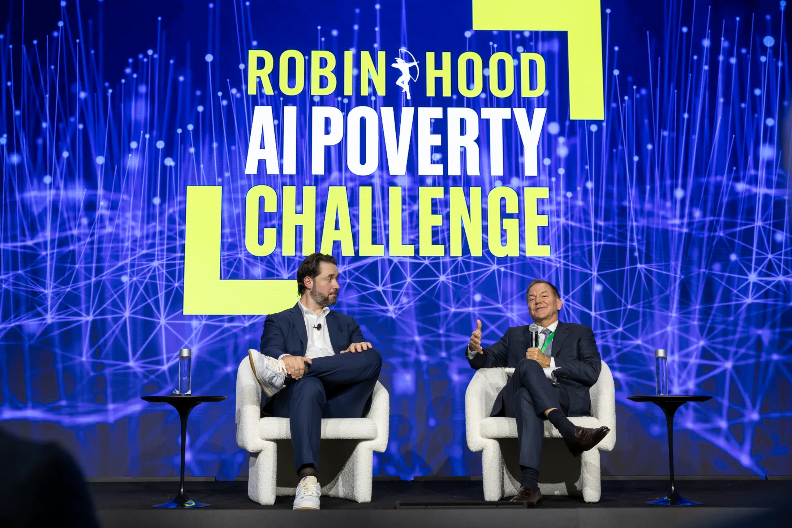 Alexis Ohanian and Pau Tudor Jones announce the Robin Hood AI Poverty Challenge on stage at a conference.