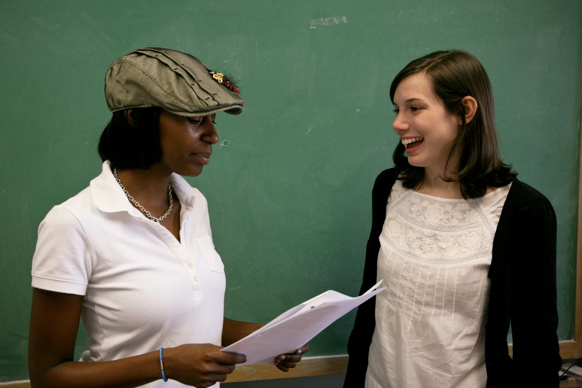 A teacher and a student discussing a paper assignment in front of a green chalk board.