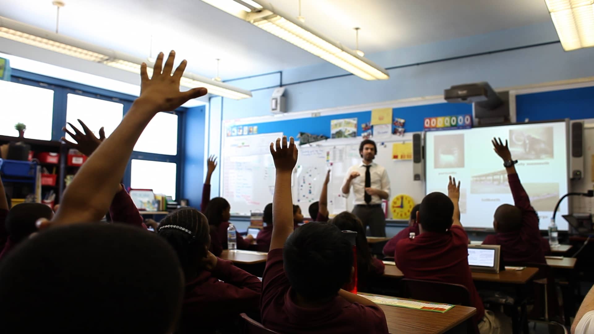 A teacher is instructing a classroom of students with their hands raised.